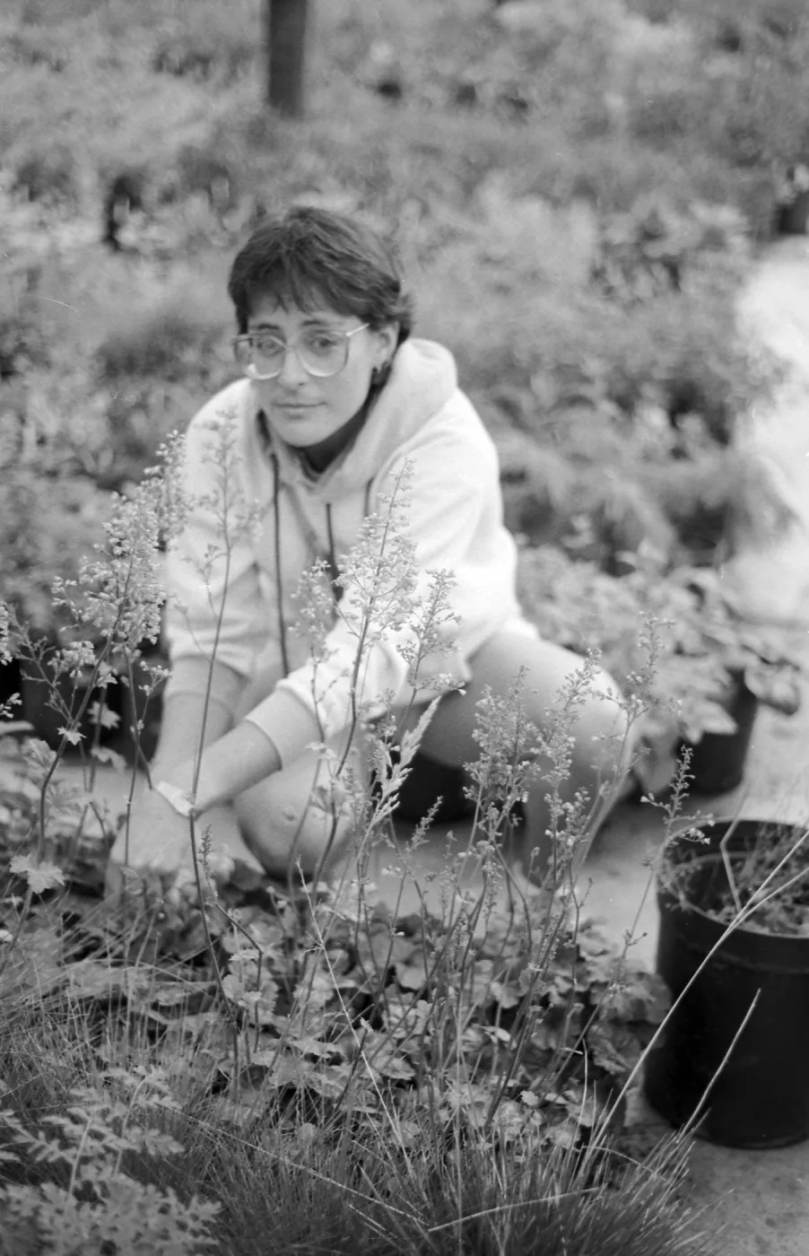 Black and white photo of a woman wearing glasses and a hoodie crouching among nursery pots, tending to plants on the ground.