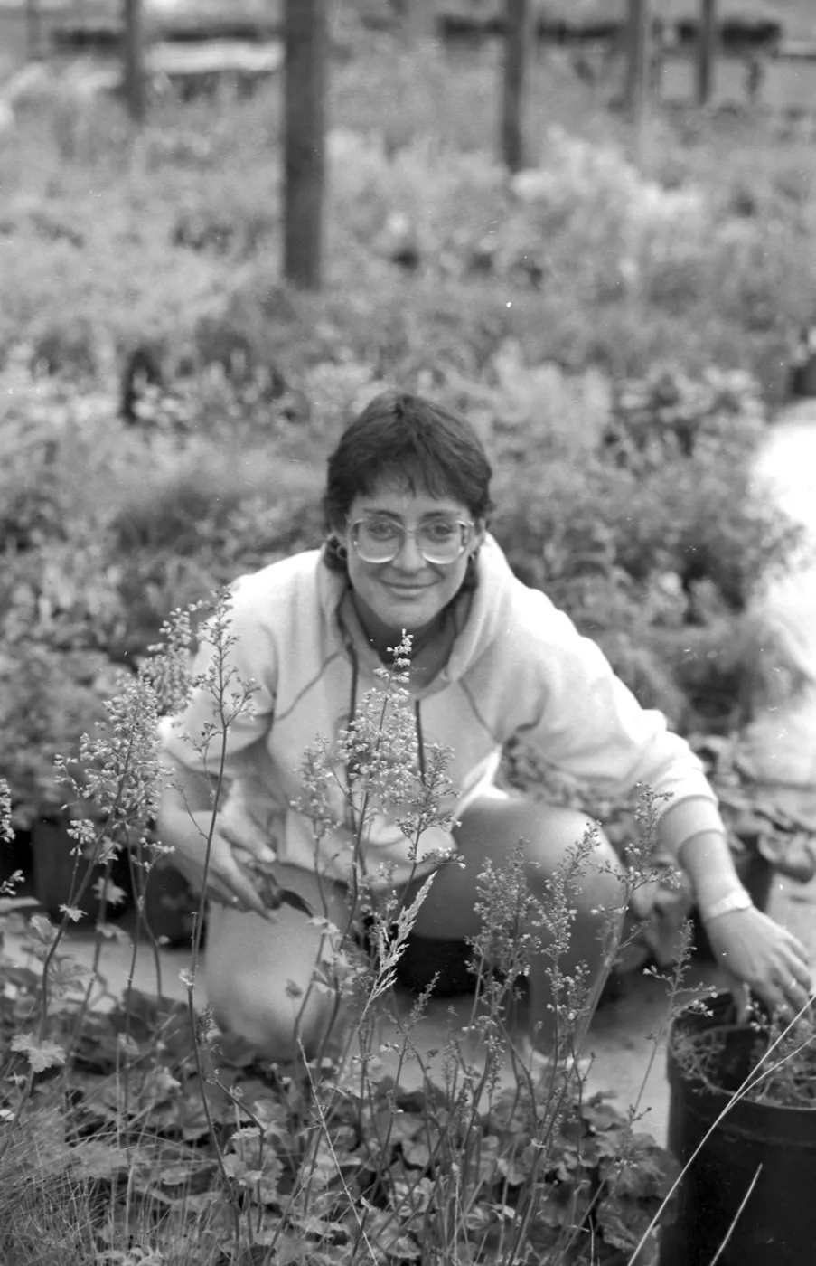 A black and white photo of a smiling woman with glasses and a hoodie squatting among nursery plants.