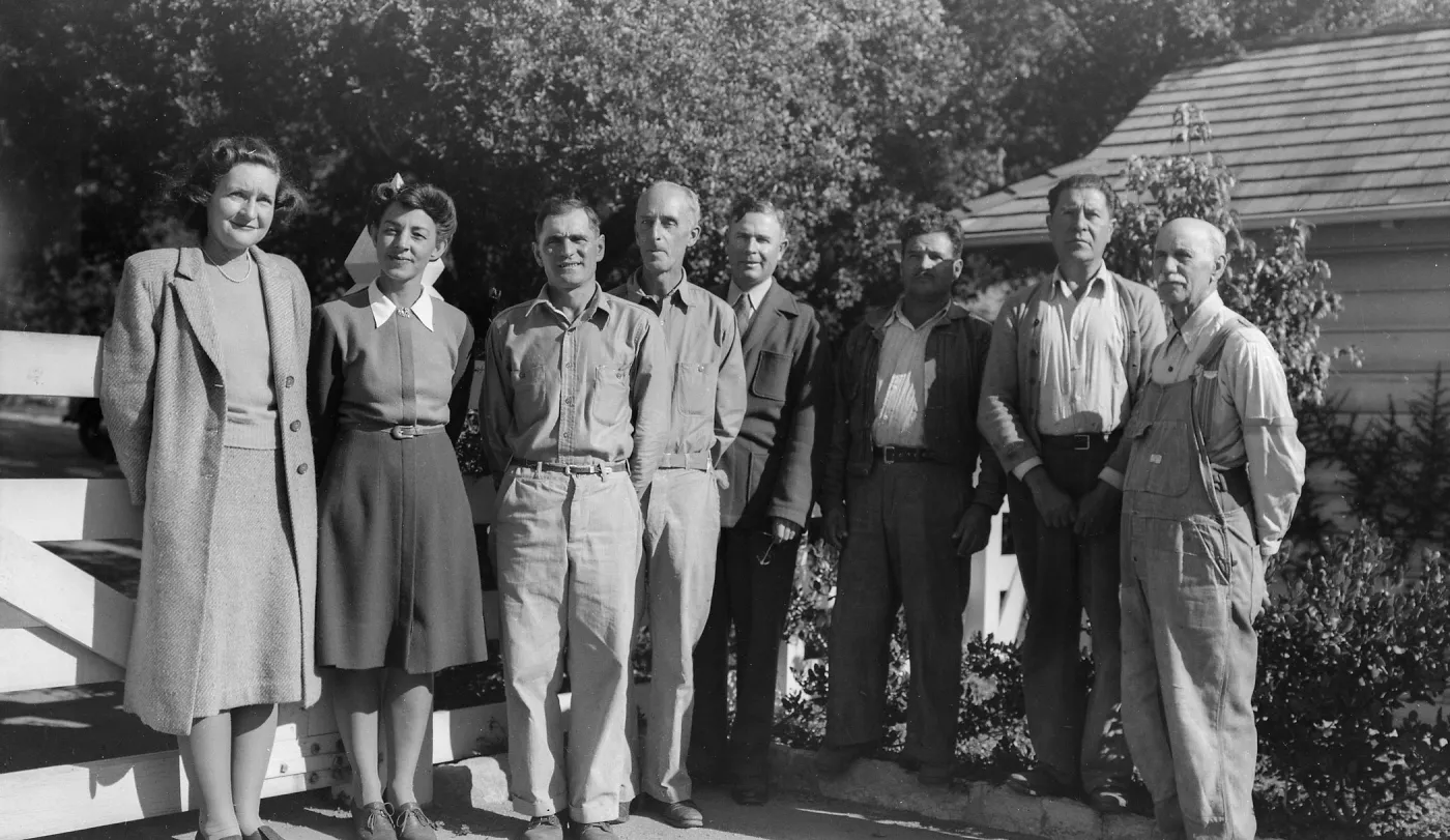 a black and white group portrait of eight adults—two women on the left and six men—standing outdoors in front of a fence and small building, posing in a line.