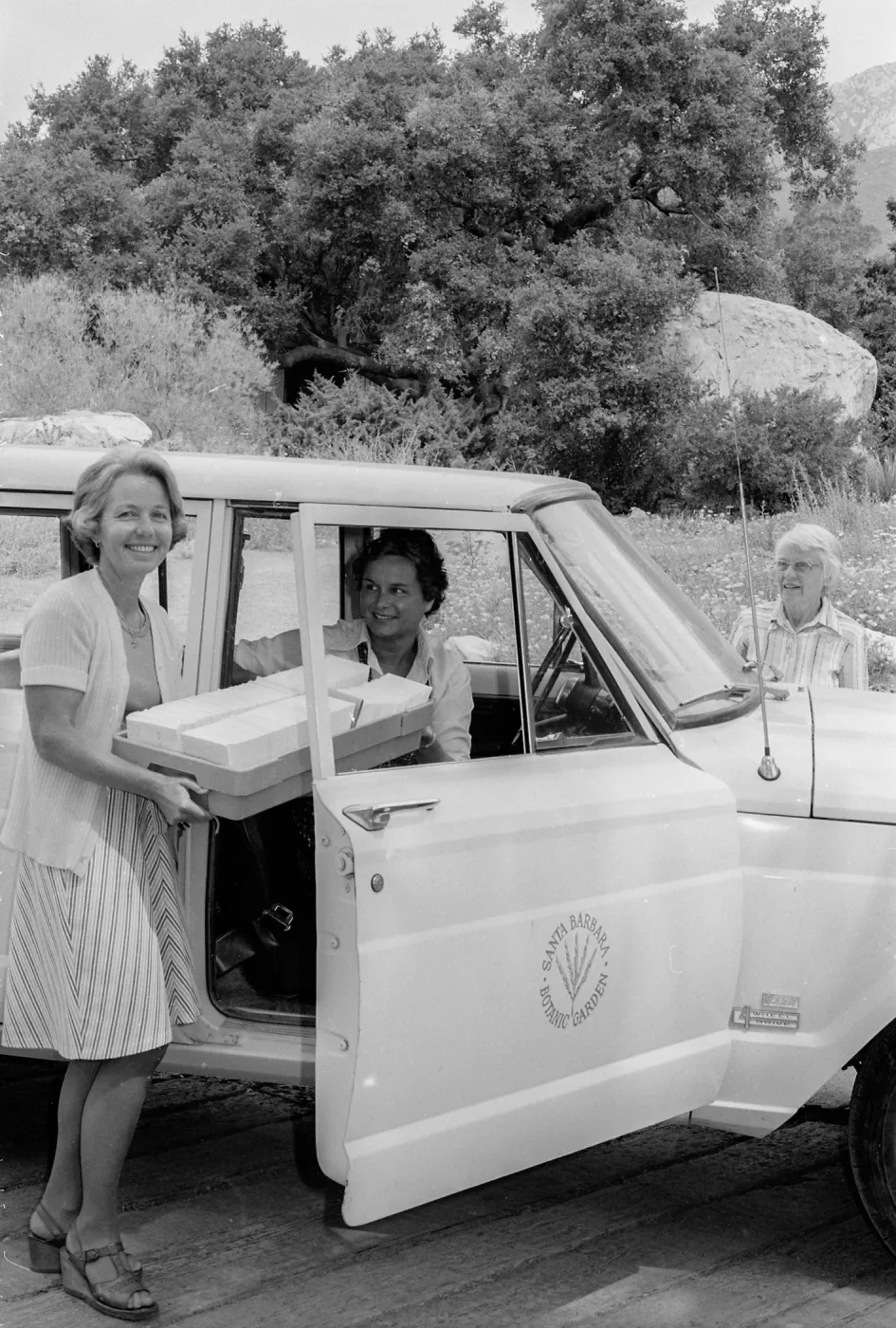 a black and white photo of three women by a parked car labeled one woman stands outside holding a cooler, another sits in the driver's seat, and an older woman stands behind the vehicle in a tree-lined area.