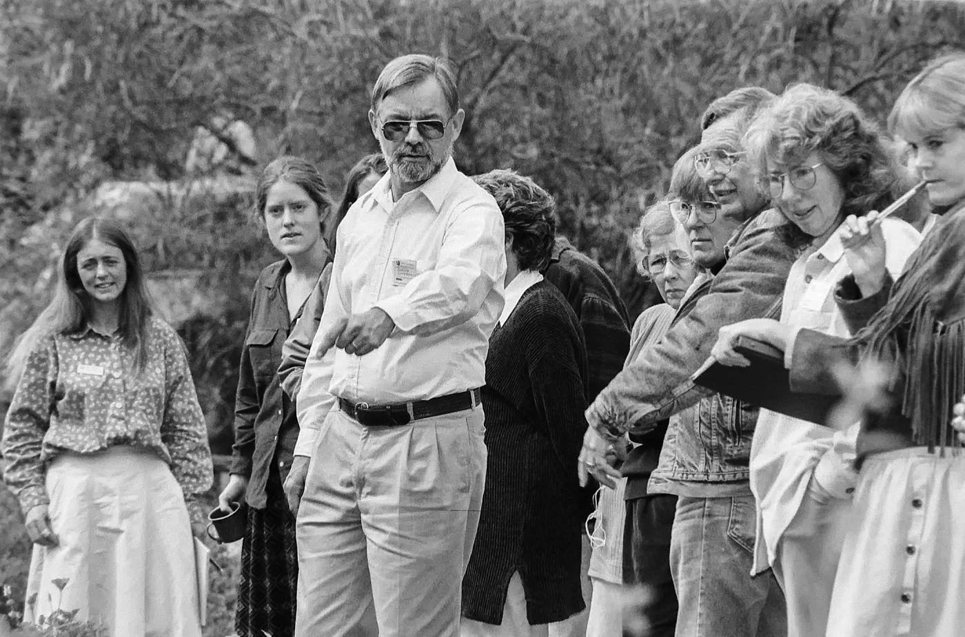 A black and white photo of a man in sunglasses pointing and speaking to a group of people outdoors who stand in a line listening.