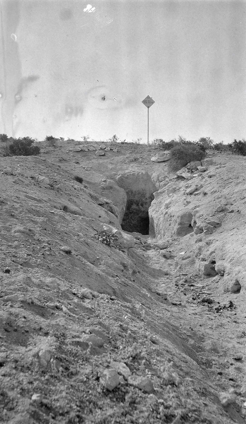 Black and white photograph of a tunnel entrance into the ground with a diamond-shaped sign on a post at the top of the hill.