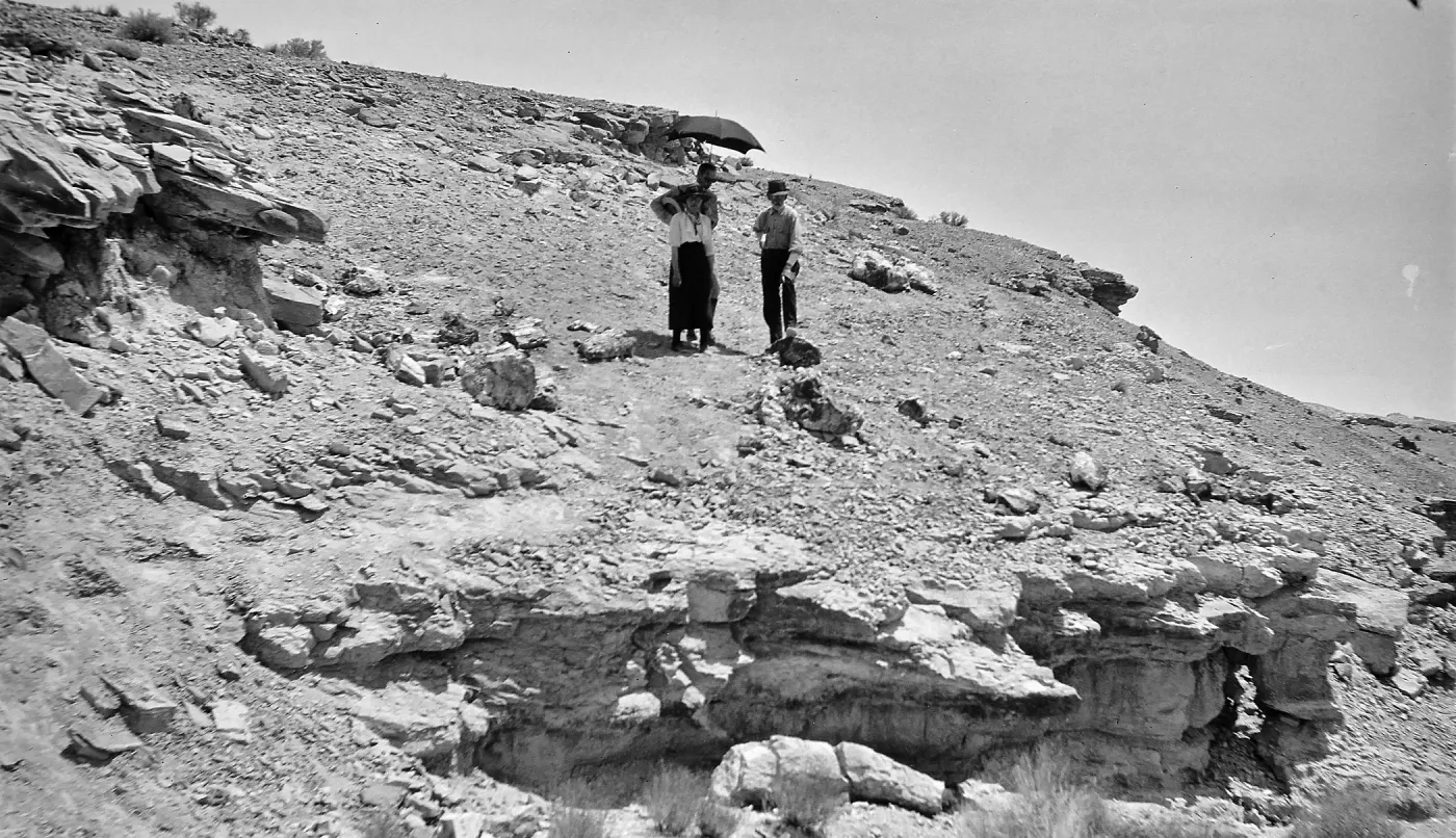 Black and white photograph of three people on a rocky hillside, two of which are underneath a black umbrella.