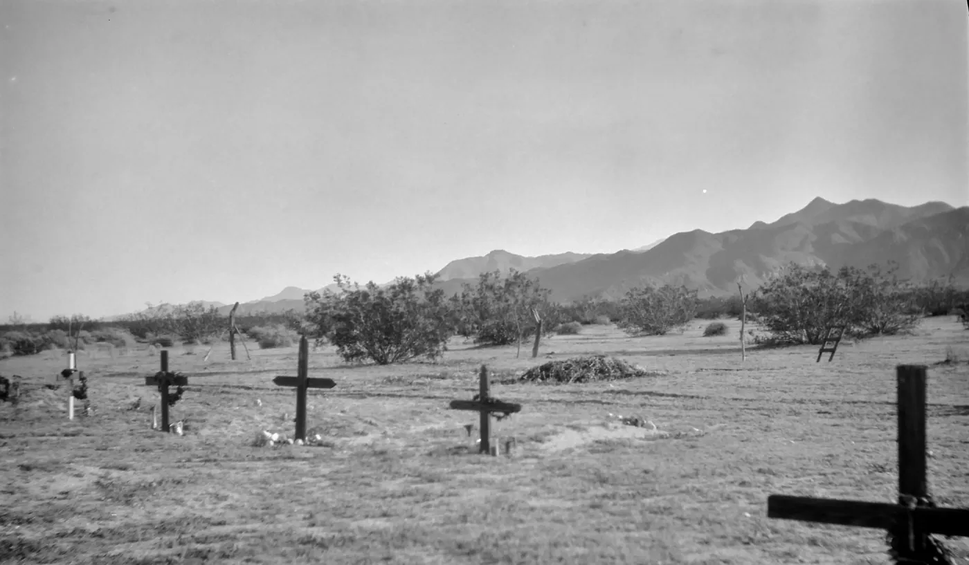 Black and white image of a graveyard with black and white crosses, with desert shrubs and hills in the background.