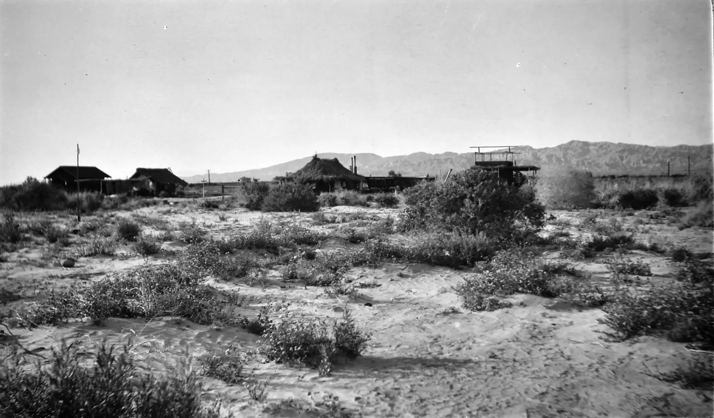 Black and white photogaph of a group of one-story buildings in a desert setting with brush and hills in the background.