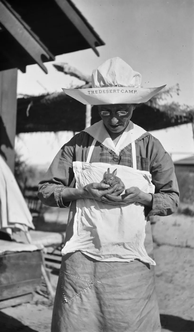 Black and white photograph of a woman in sunglasses and a large white hat reading The Desert Camp holding a small rabbit in her hands.