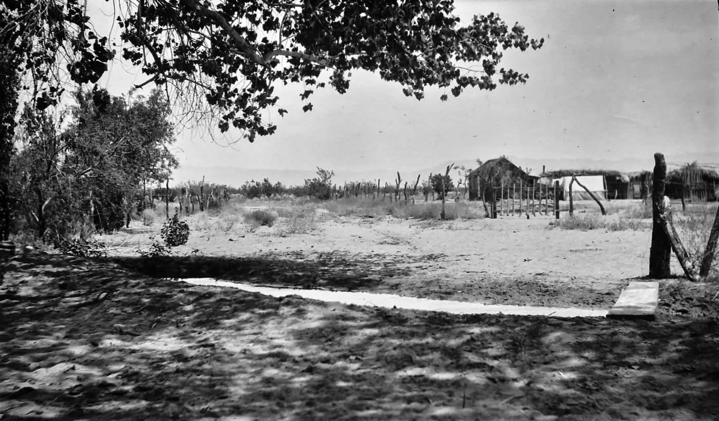 Black and white photograph of an irrigation channel in the foreground and a vineyard, wooden structure, and hills in the background.