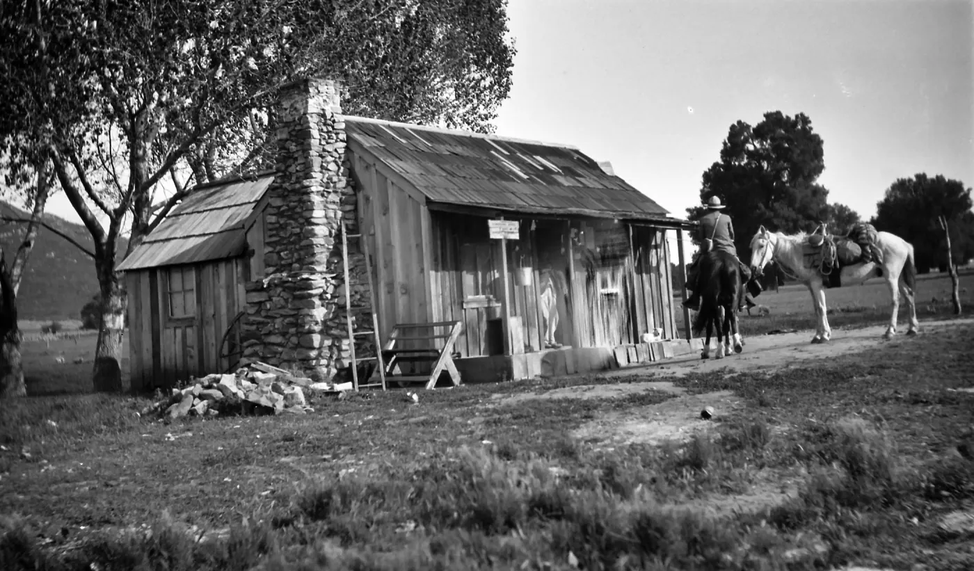 Black and white photograph of a small wooden house with a stone chimney and a man on horseback in front, a white horse next to him and trees in the distance.