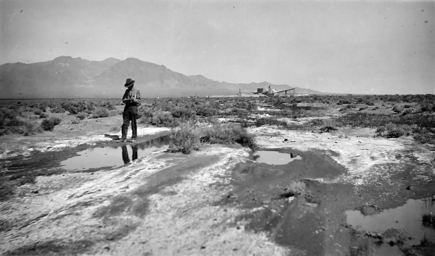 Black and white photograph of a sandy field covered in puddles, with a man in a hat in the foreground and a wooden structure and hills in the background.