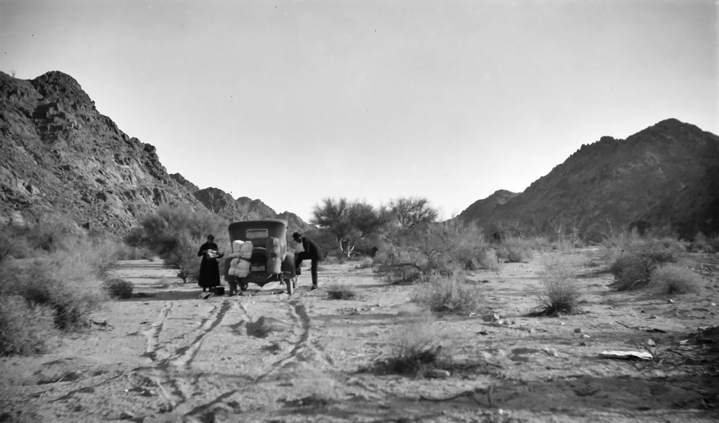 Black and white photograph of a black car and a man and woman in a desert landscape with scrub and low hills around them. The man has his foot propped on the car and the woman is looking at something in her hands.
