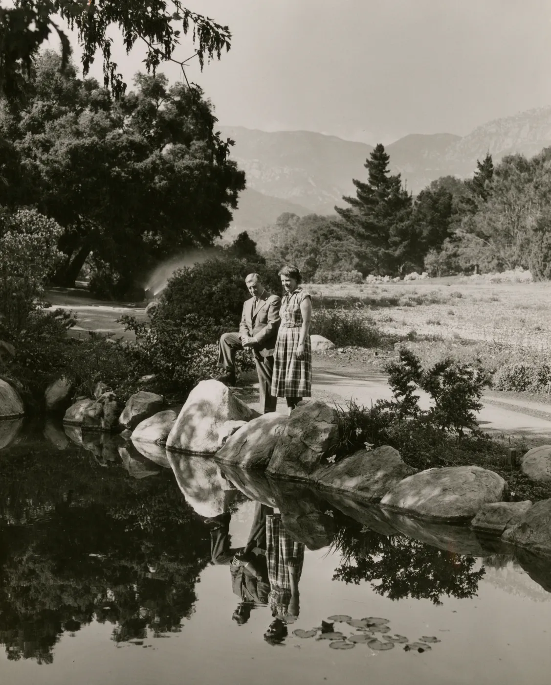 Black and white photo of a man in a suit and a woman in a plaid dress look out over a pond lined with large rocks, the man has a leg propped up on one of the rocks. 