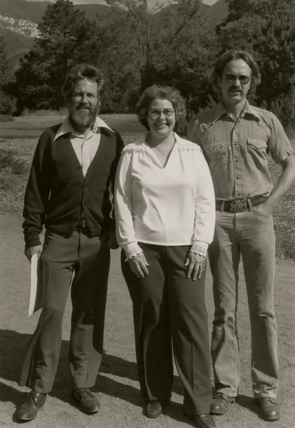A black and white photo of two men and a woman, smiling with windswept hair, in a rocky field at the edge of a copse of trees. 