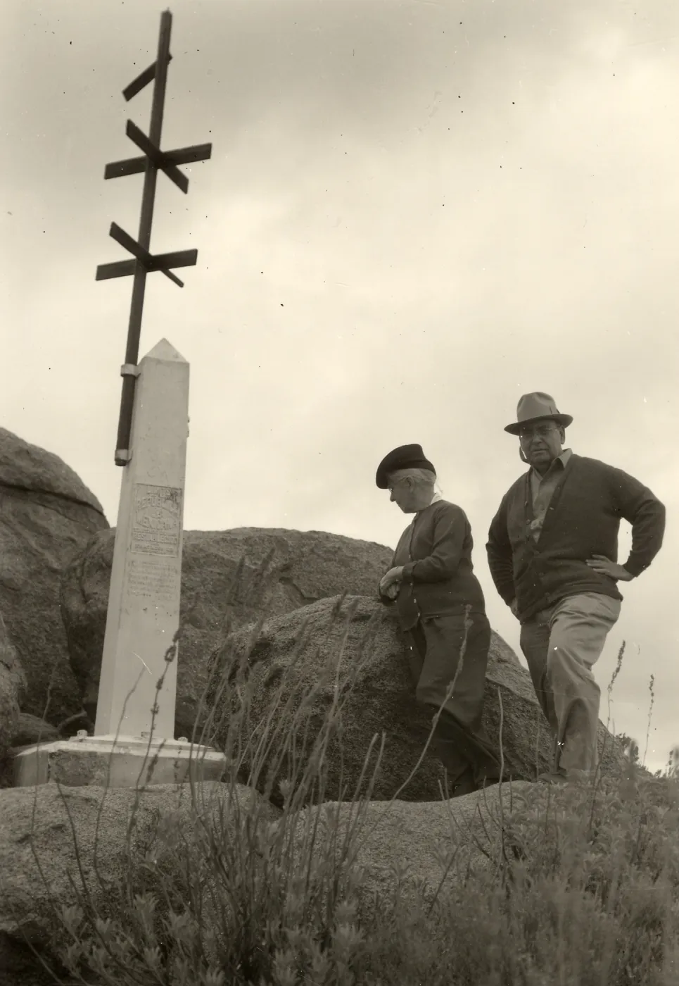 Black and white photograph of two people standing to the right of a monument on large rocks.