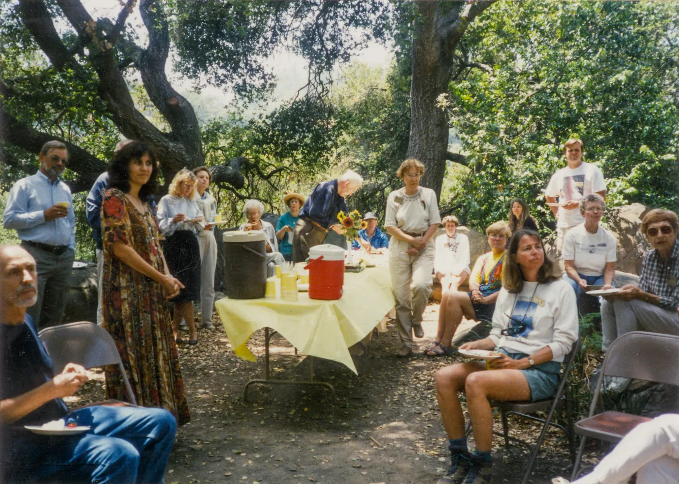 Group of people sitting and standing around table of food in woods looking to the right. 