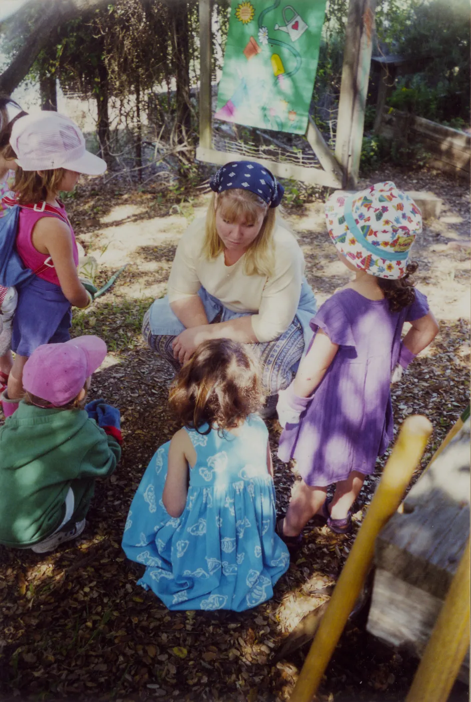 Woman talking to children in wood clearing with colorful panel of watering plants in background. 