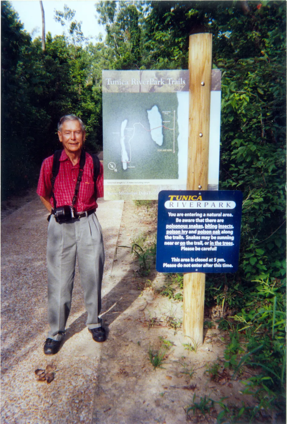 Photograph of male wearing red shirt and camera standing at trail sign with lush green background. 