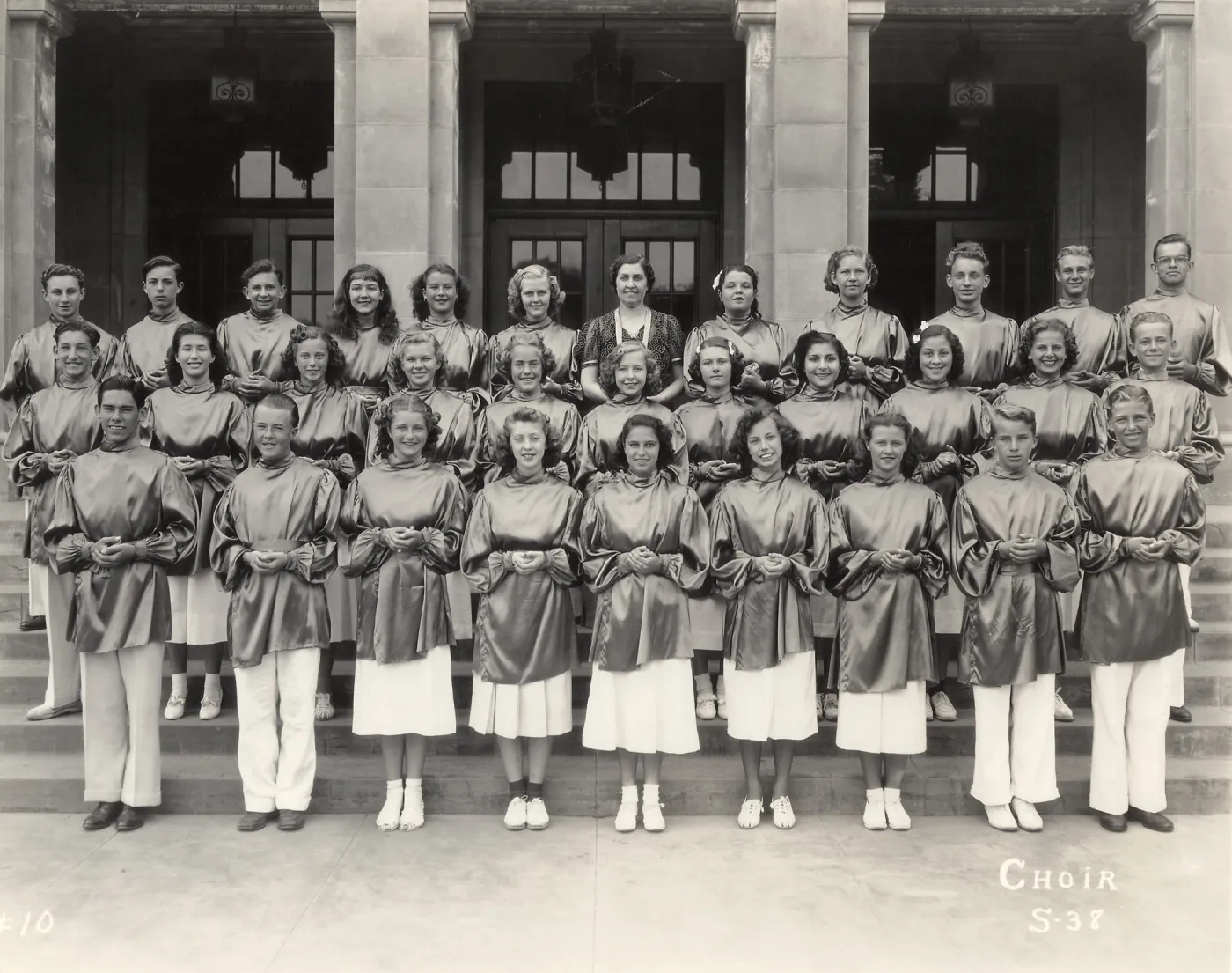 Black and white photograph of high school choir; three rows of people in cloaks stand in front of building.