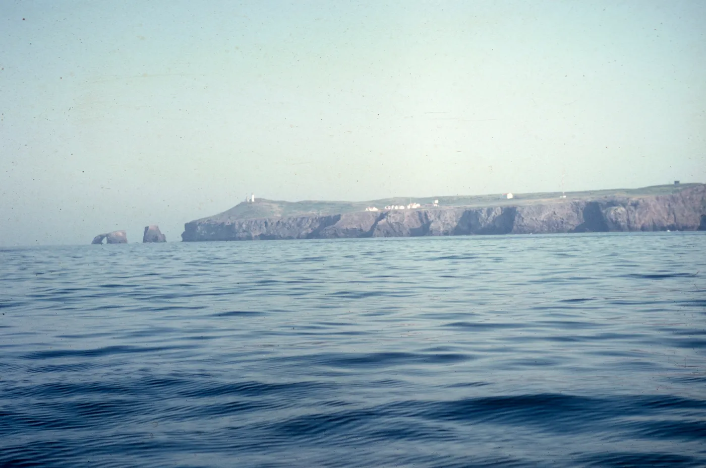 An island edged by rocky cliffs. Two sea stacks are visible off the end of the island, the furthest of which forms an arch. Several white building are visible at the top of the cliff, with one, taller while building alone at the top of the headland. 