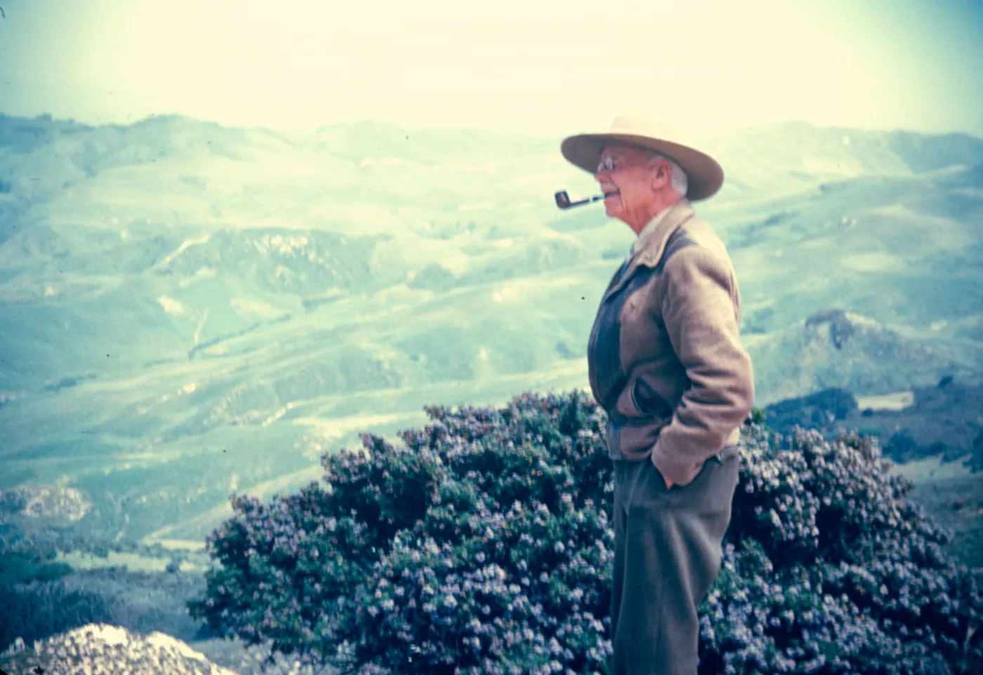 An older man in a straw had with a pipe in his mouth looks out over a landscape of mountains and valleys. 