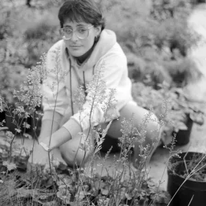 Black and white photo of a woman wearing glasses and a hoodie crouching among nursery pots, tending to plants on the ground.
