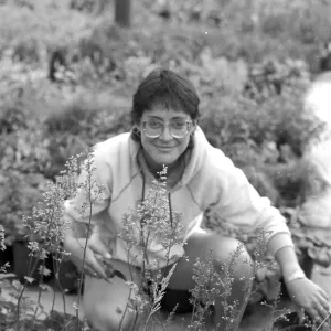 A black and white photo of a smiling woman with glasses and a hoodie squatting among nursery plants.
