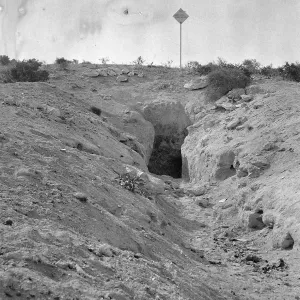 Black and white photograph of a tunnel entrance into the ground with a diamond-shaped sign on a post at the top of the hill.
