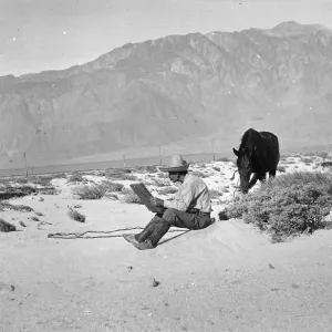 Black and white photo of a man in a hat sitting and sketching in a large book with a black horse next to him and a mountain range behind him.