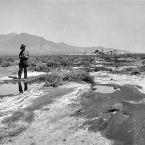 Black and white photograph of a sandy field covered in puddles, with a man in a hat in the foreground and a wooden structure and hills in the background.