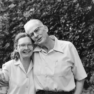 Black and white photograph of a man and woman against a background of bushes. Both are wearing glasses and white shirts and smiling at the camera. 