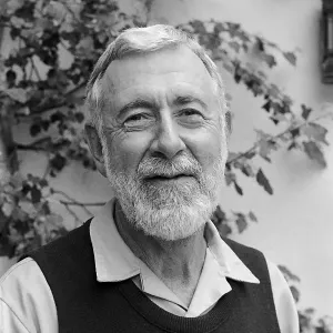 Close-up portrait of an older smiling man with a beard and sweater vest against a leafy wall background.