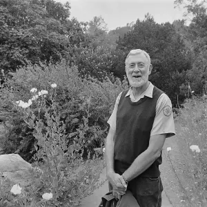 Black-and-white photo of an older man in a vest smiling and standing on a garden path surrounded by wildflowers, shrubs, and trees.
