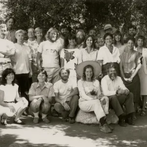 A black and white photo of a group of 26 smiling people posed on the edge of a path. Five people crouch or sit on a large rock, the rest stand in two rows behind them.