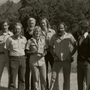 A black and white photo of seven people wearing slacks and button-up shirts stand in a rocky field at the edge of a copse of trees.