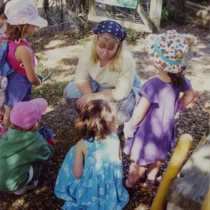 Woman talking to children in wood clearing with colorful panel of watering plants in background. 