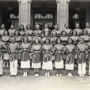 Black and white photograph of high school choir; three rows of people in cloaks stand in front of building.