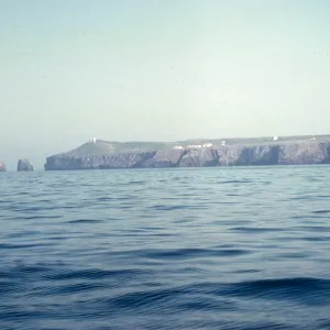 An island edged by rocky cliffs. Two sea stacks are visible off the end of the island, the furthest of which forms an arch. Several white building are visible at the top of the cliff, with one, taller while building alone at the top of the headland. 