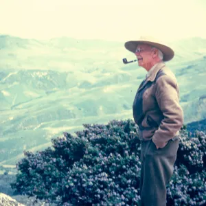 An older man in a straw had with a pipe in his mouth looks out over a landscape of mountains and valleys. 