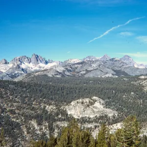 An evergreen forest grows at the foot of and between a range of sharp, grey, snowy mountains. 