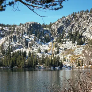 Pine trees grow on the far side of a lake at the base of a snow-covered mountain ridge.
