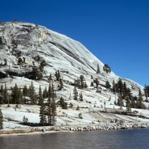 A smooth, rounded, white and grey stoned mountain dotted with pine trees on the far side of a calm lake. 