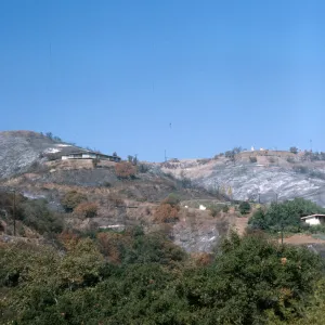 A few low, white houses on rounded mountain slopes among brown, burned vegetation.