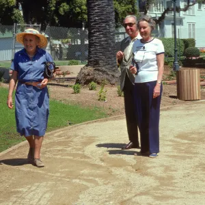 Three older people, a woman in a blue dress and straw hat, a woman in a white shirt and navy slacks, and a man in am olive suit and sunglasses, smile on a path between grass fields and large palm trees. 