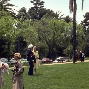 Two older people, one in a yellow dress and one in navy blue suit talk in a grassy yard adjacent to a road lined with old model cars. A woman in a tan trench coat with a camera talks to someone out of frame. 