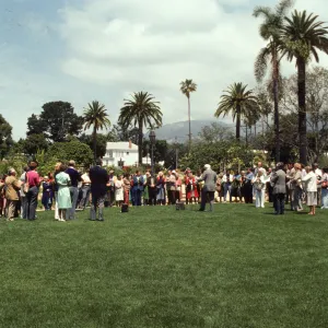 A large group of well-dressed people stand in a semicircle in a grassy yard. An older man in a grey suit stands in the center of the semi-circle.