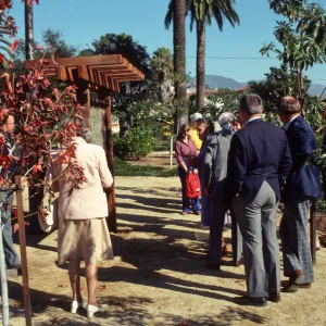 A group of well-dressed people gather in a garden around a large, wooden exhibit sign containing a map. 