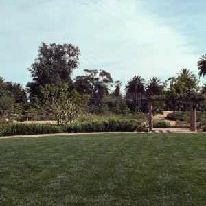 A uniform, grassy yard bordered by a walking path, several trees and bushes, and two benches beneath a pergola. 