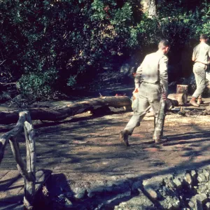 Two men in khaki work clothes chopping wood on muddy, stony ground at the edge of a wooded area. A rustic wooden bridge leads into the area.
